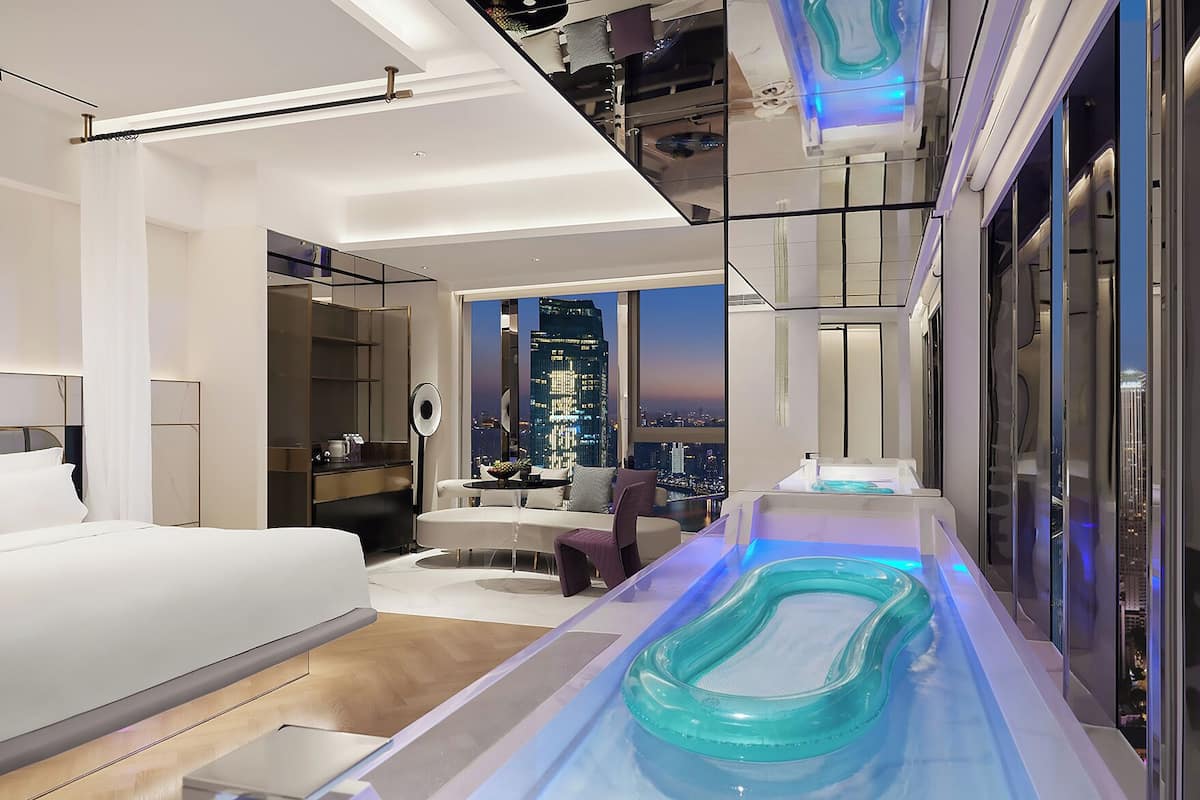 Ein modernes Hotelzimmer mit Bett, einer Sitzecke, einem kleinen Tisch mit Obstschale, einem großen Fenster mit Stadtblick und einer integrierten Whirlwanne.