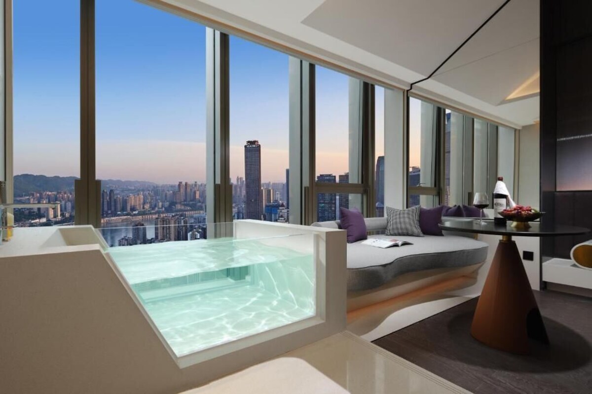 Een moderne hotelkamer met een groot stadsgezicht, een bubbelbad in een glazen ruimte, en een gezellige zithoek met een tafel en stoelen.