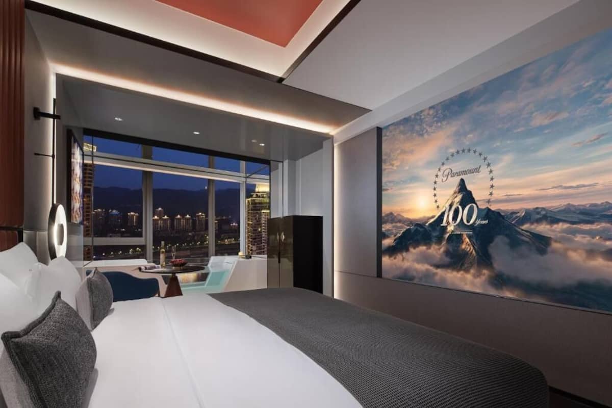 Ein modernes Hotelzimmer mit einem großen Bett, Blick auf die nächtliche Stadt und einem Kunstwerk mit Bergmotiv an der Wand.