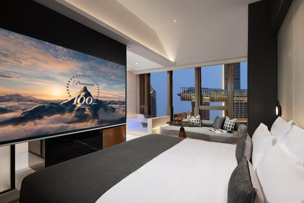 Una camera d'albergo moderna con una grande televisione a schermo piatto che mostra una scena montana, un letto grande, un'area salotto con vista sulla città e un balcone con ringhiera.