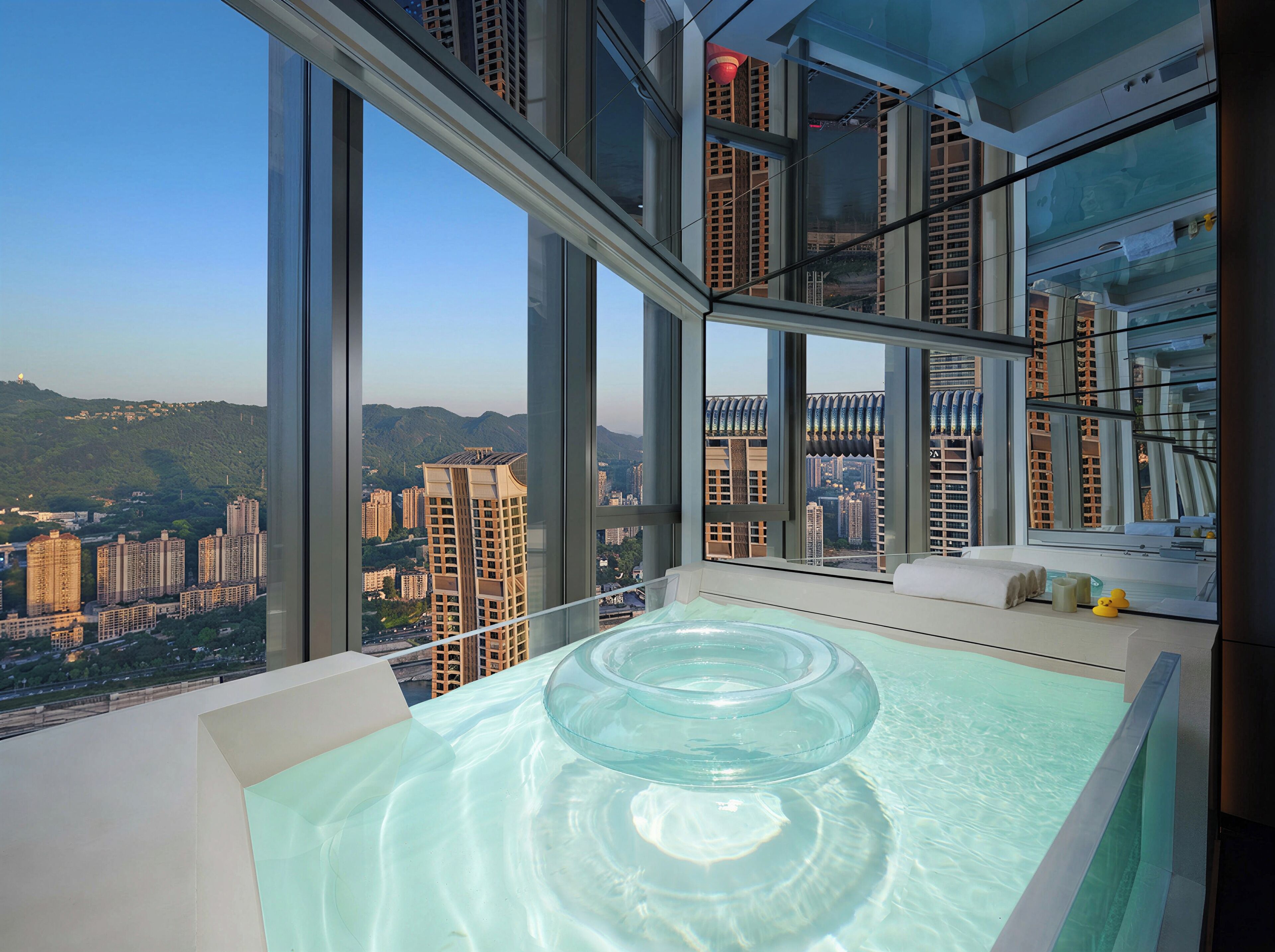 A high-rise hotel room with a large window, a glass bathtub, and a view of the cityscape.
