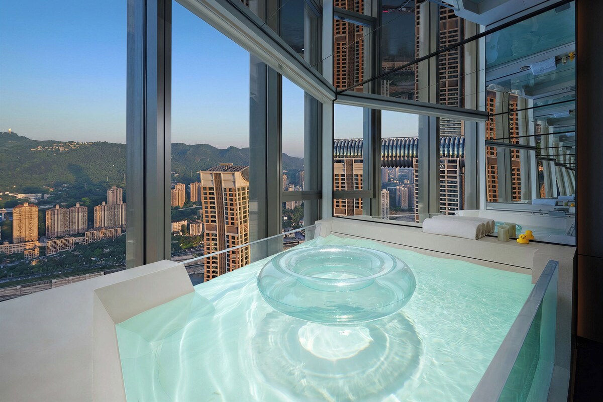 Hotelkamer hoog in een wolkenkrabber met een groot raam, een glazen ligbad en uitzicht op de stad.