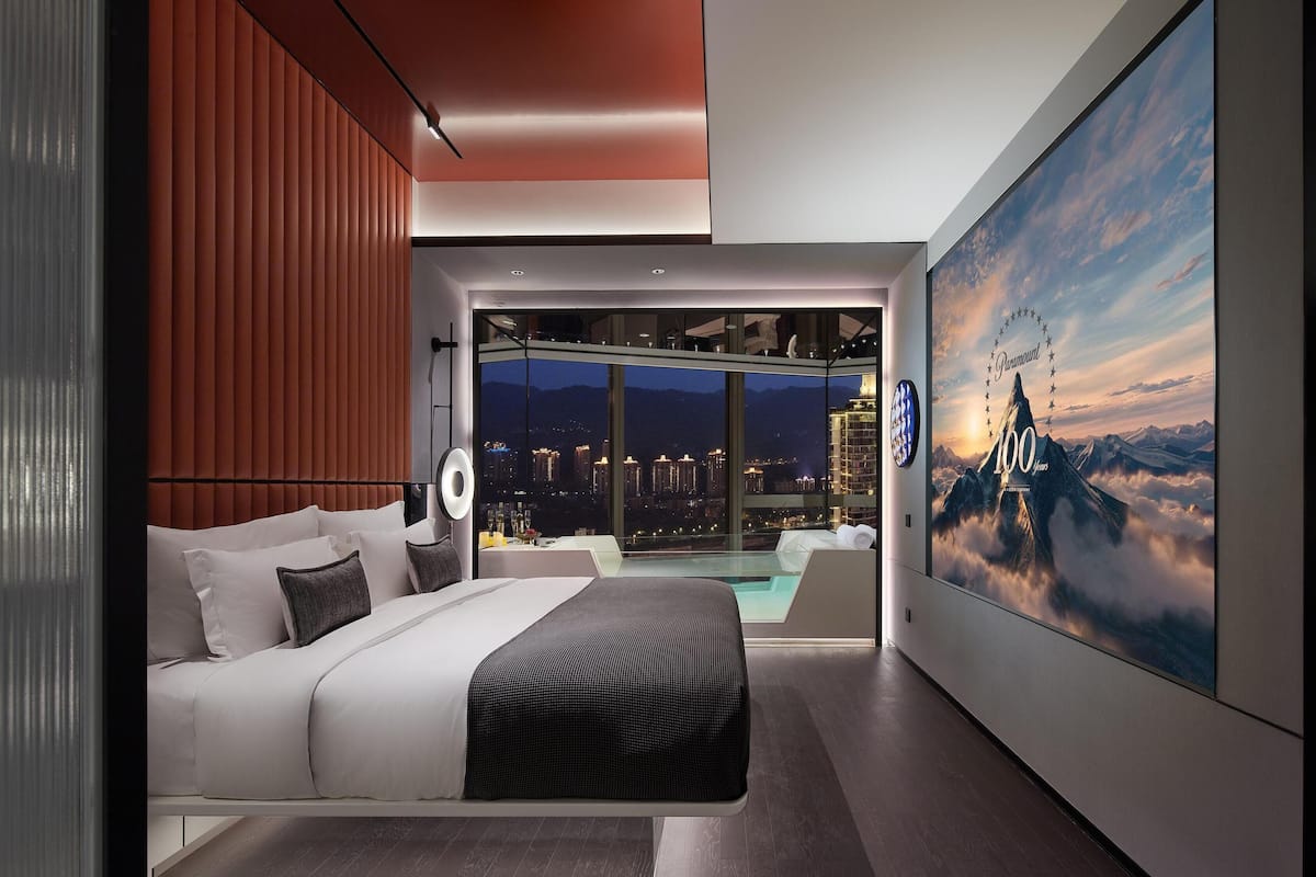 Ein modernes Schlafzimmer mit einem großen Bett, einer Glaswand, Blick auf die beleuchtete Stadt und einem großen Poster an der Wand.