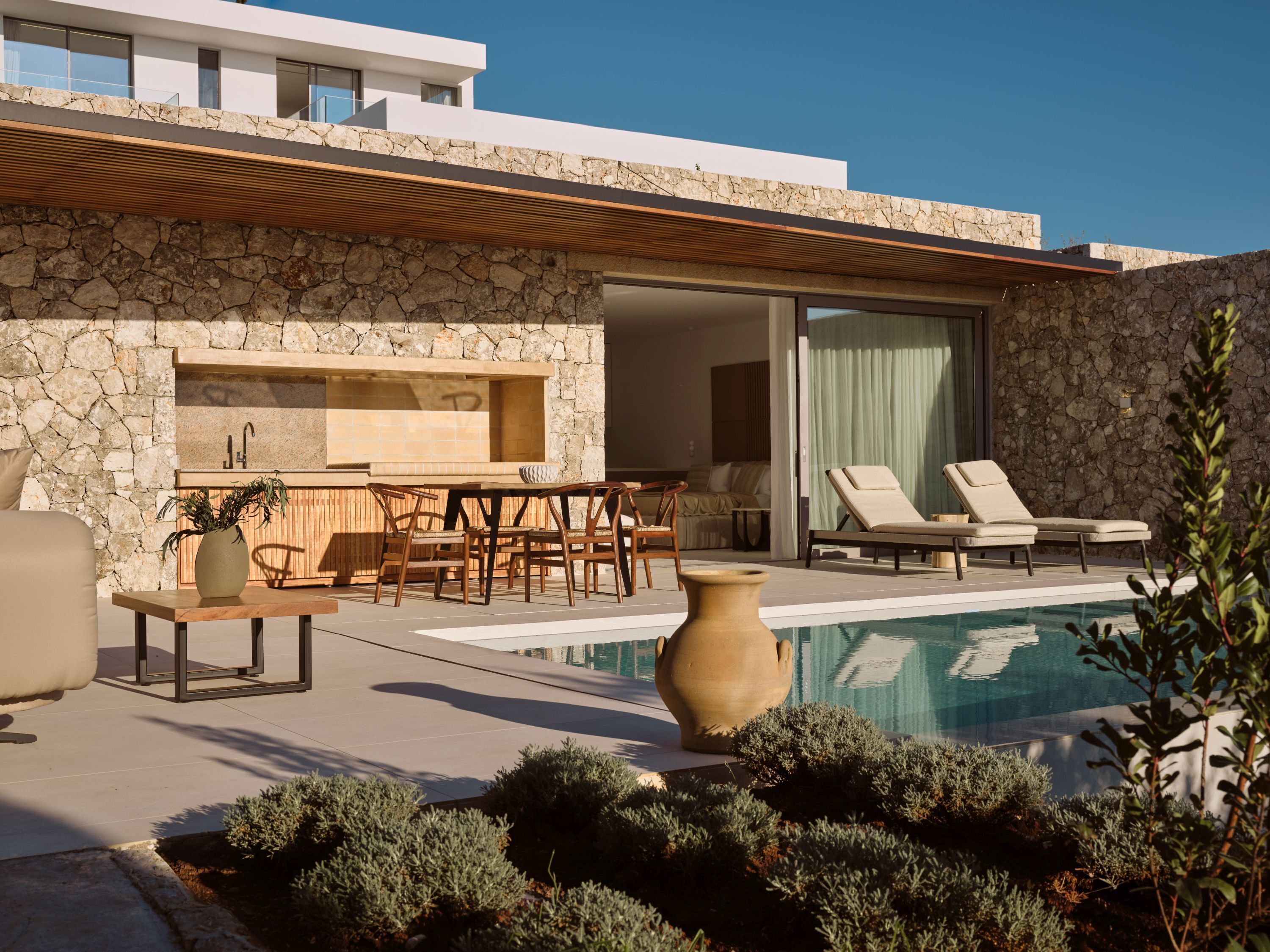 Un área de piscina moderna al aire libre con muros de piedra, una pérgola de madera y sillones de descanso.