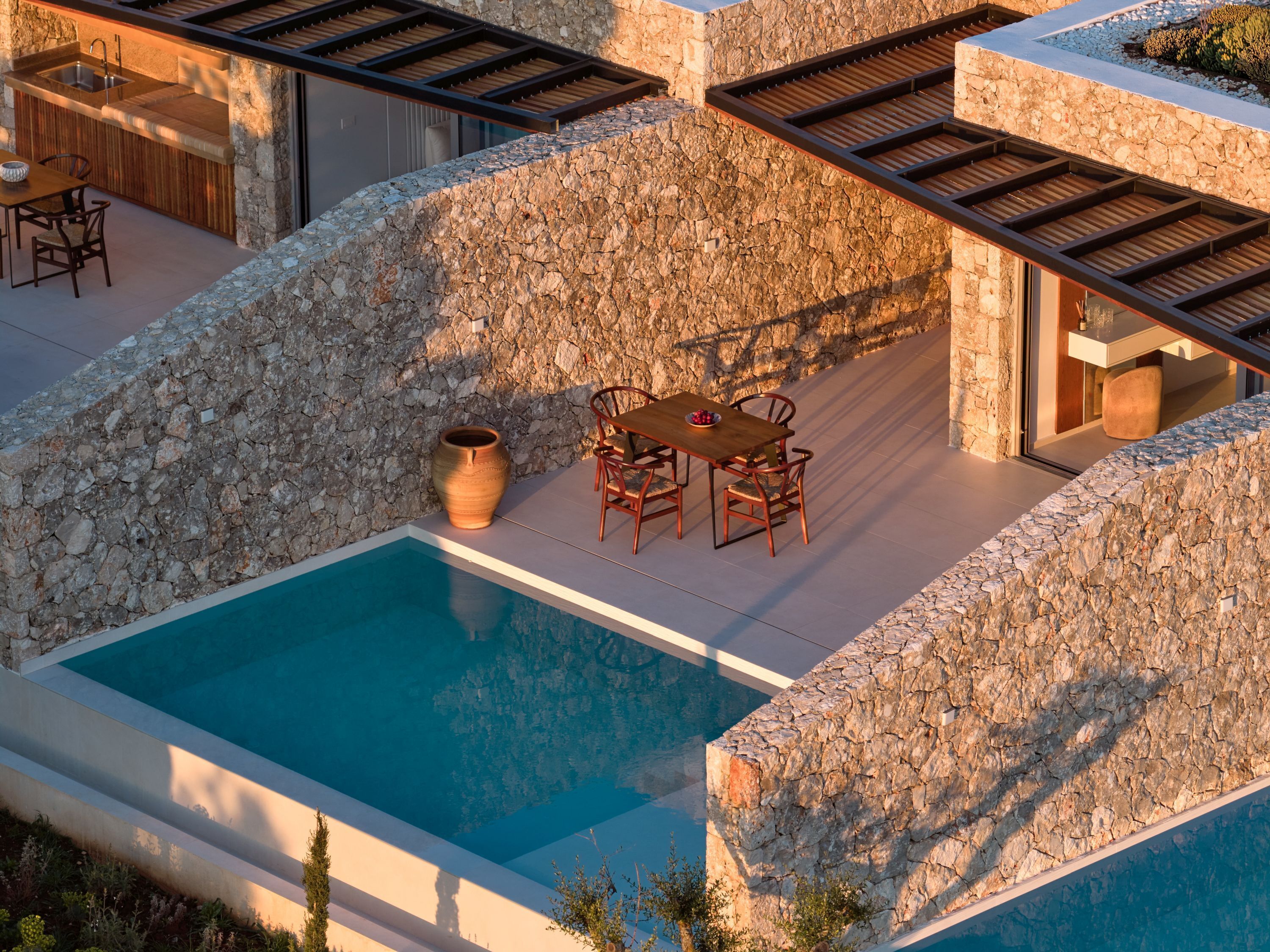 A poolside area with a stone wall, a wooden table with chairs, and a large vase.