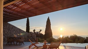 Una terrazza esterna con un muro in pietra, una pergola in legno e vista sul tramonto sul mare.