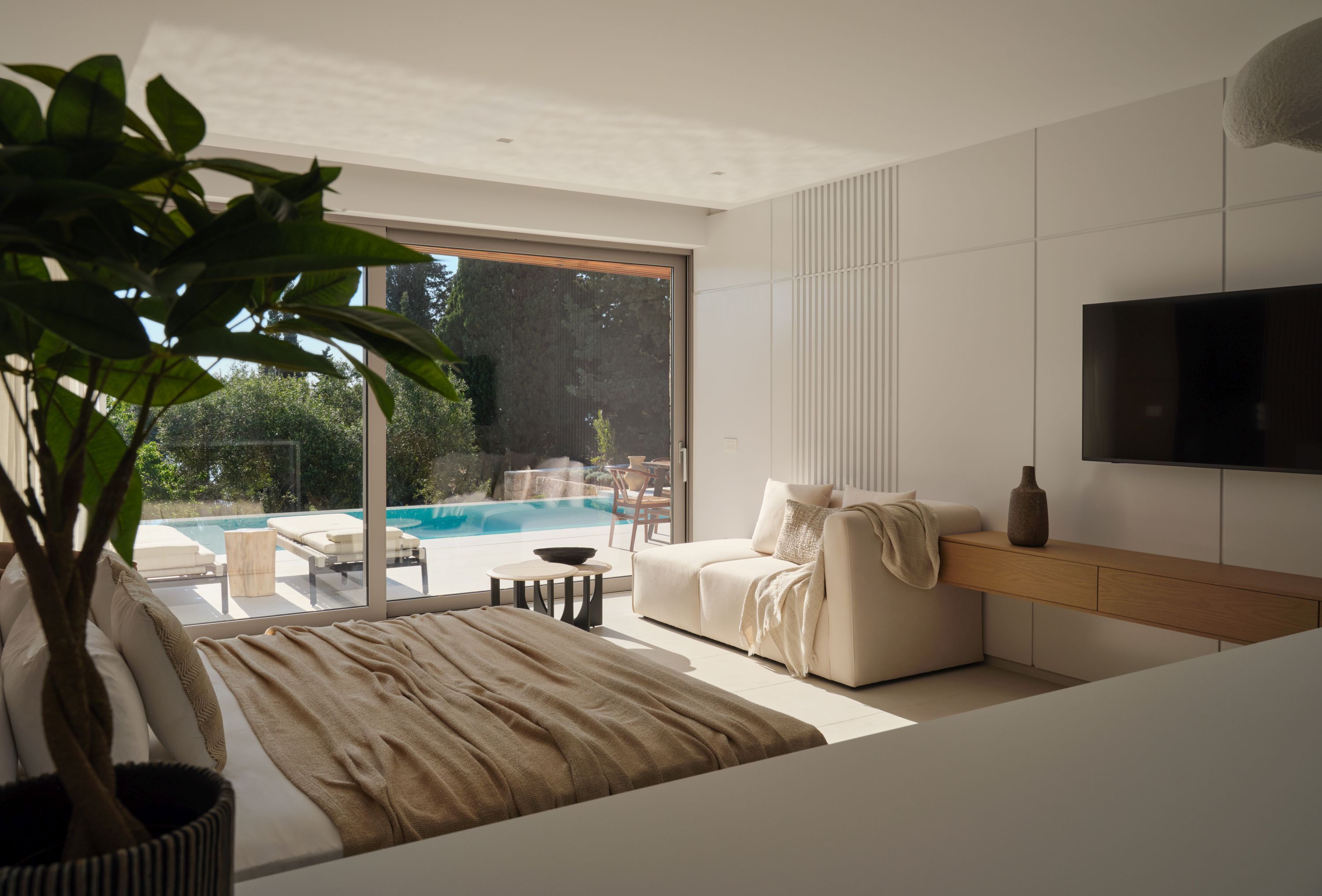 A modern living room with a large sliding glass door, a beige sofa, a TV mounted on the wall, and a potted plant.