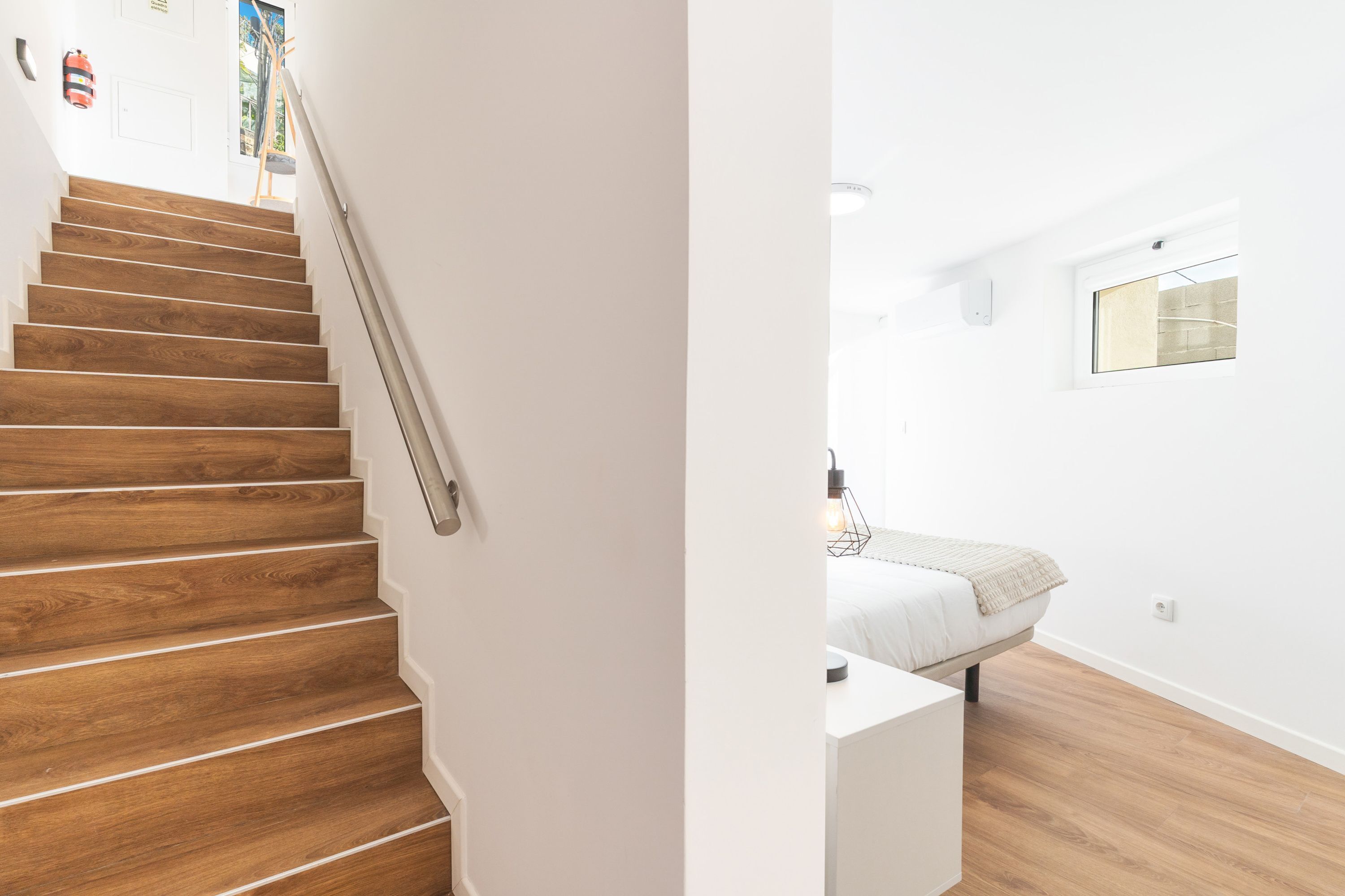 A staircase with wooden steps and a handrail, leading to a room with a bed, a small table, and a window.
