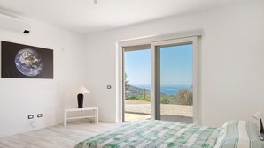 A bedroom with a bed, a nightstand, a lamp, a painting of Earth, and a sliding glass door leading to a balcony with a view of the sea. - Adriana Trilocale Apartment with Sea View and Wi-Fi (Lacona)