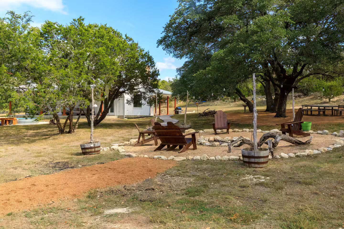 Un área de estar al aire libre con bancos y mesas de madera, rodeada de árboles y un edificio pequeño al fondo.