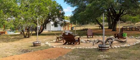 Un área de estar al aire libre con bancos y mesas de madera, rodeada de árboles y un edificio pequeño al fondo.