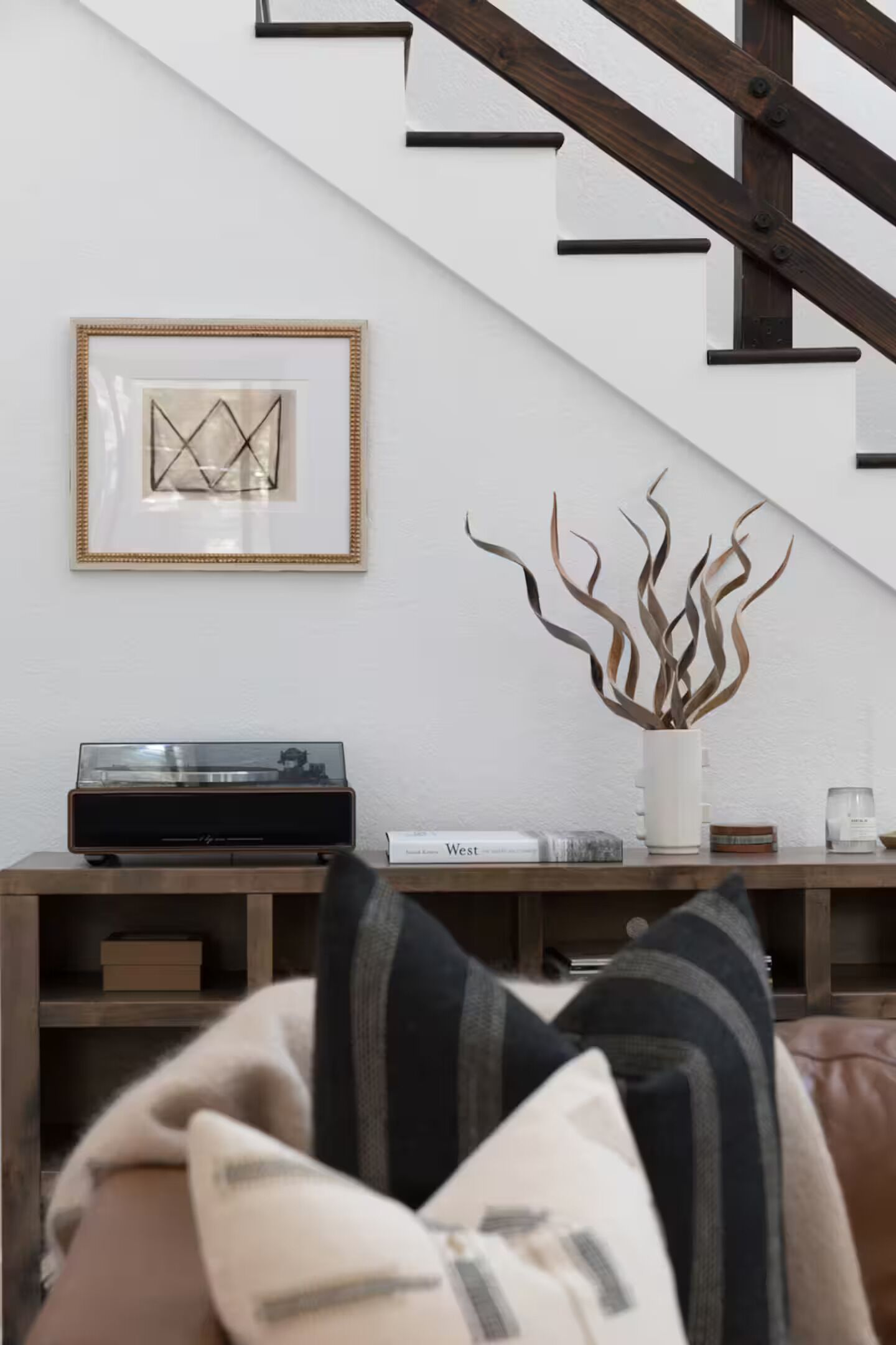 A living room with a staircase, a framed picture on the wall, a plant in a white vase, and a record player on a wooden console table.