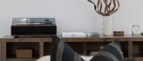 A living room with a staircase, a framed picture on the wall, a plant in a white vase, and a record player on a wooden console table.