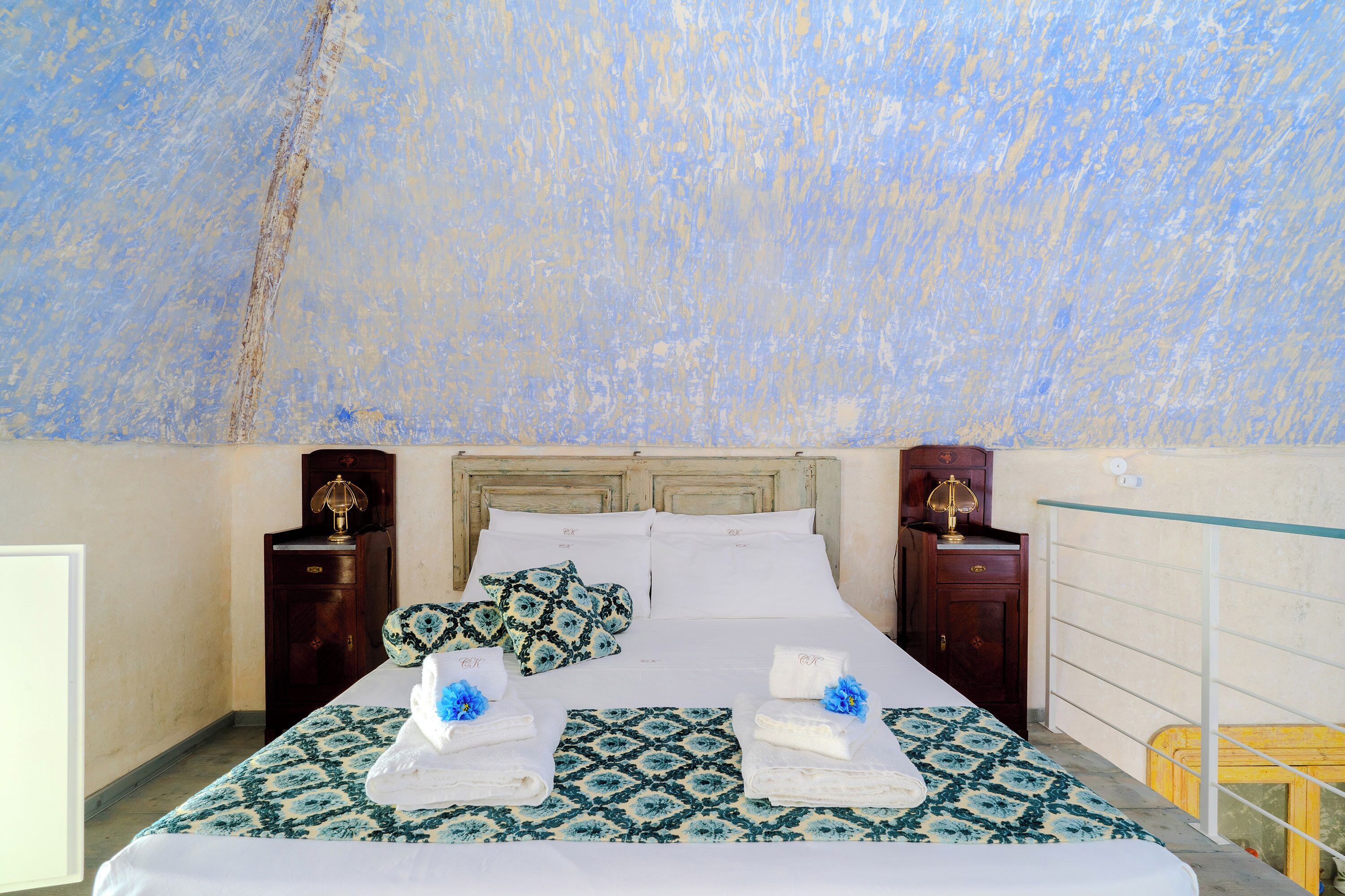 Una cama bien hecha con una colcha estampada, dos mesitas de noche con lámparas y una pared con textura.