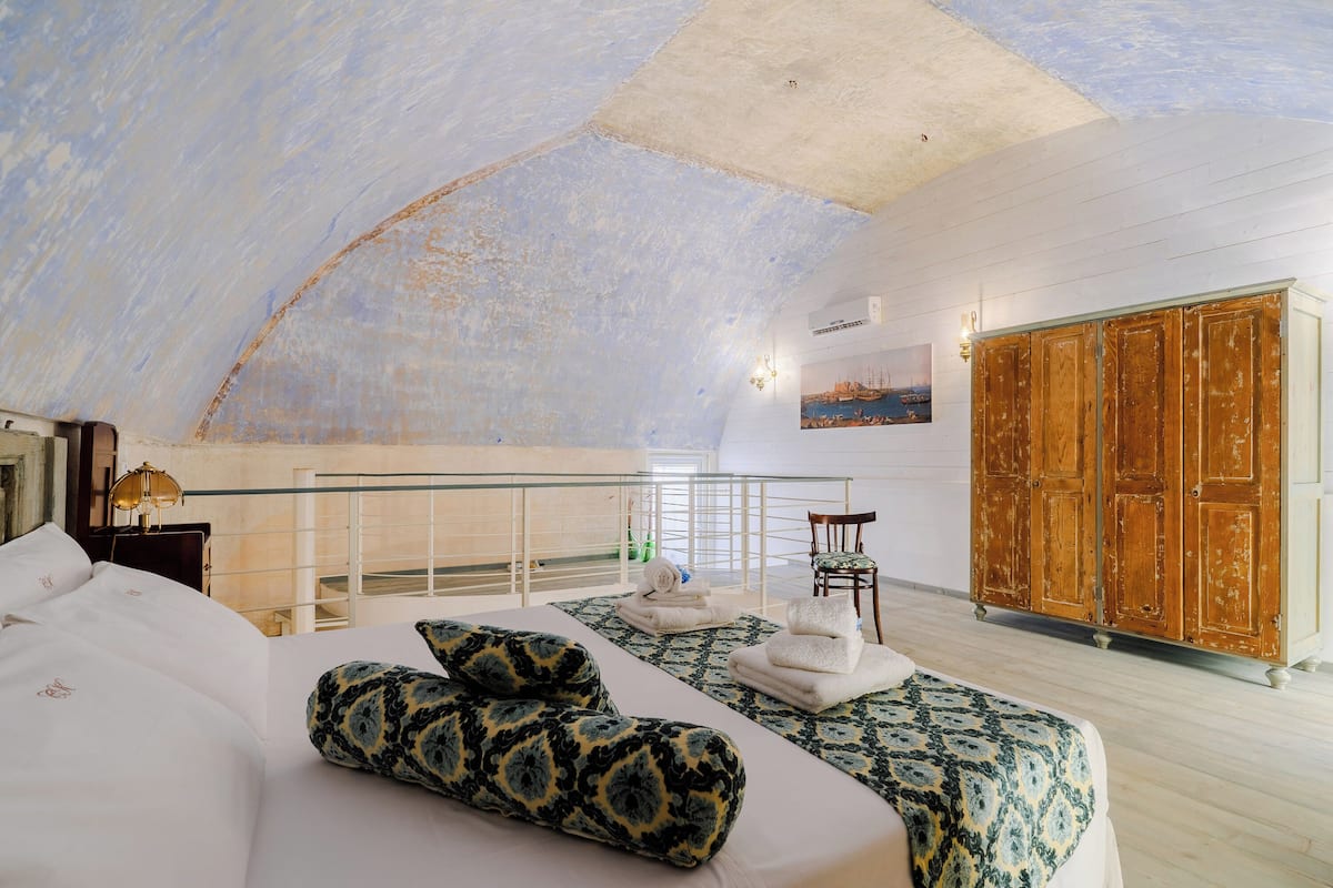A spacious room with a high ceiling, a bed with patterned pillows, a wooden wardrobe, and a view of the sea through a balcony.