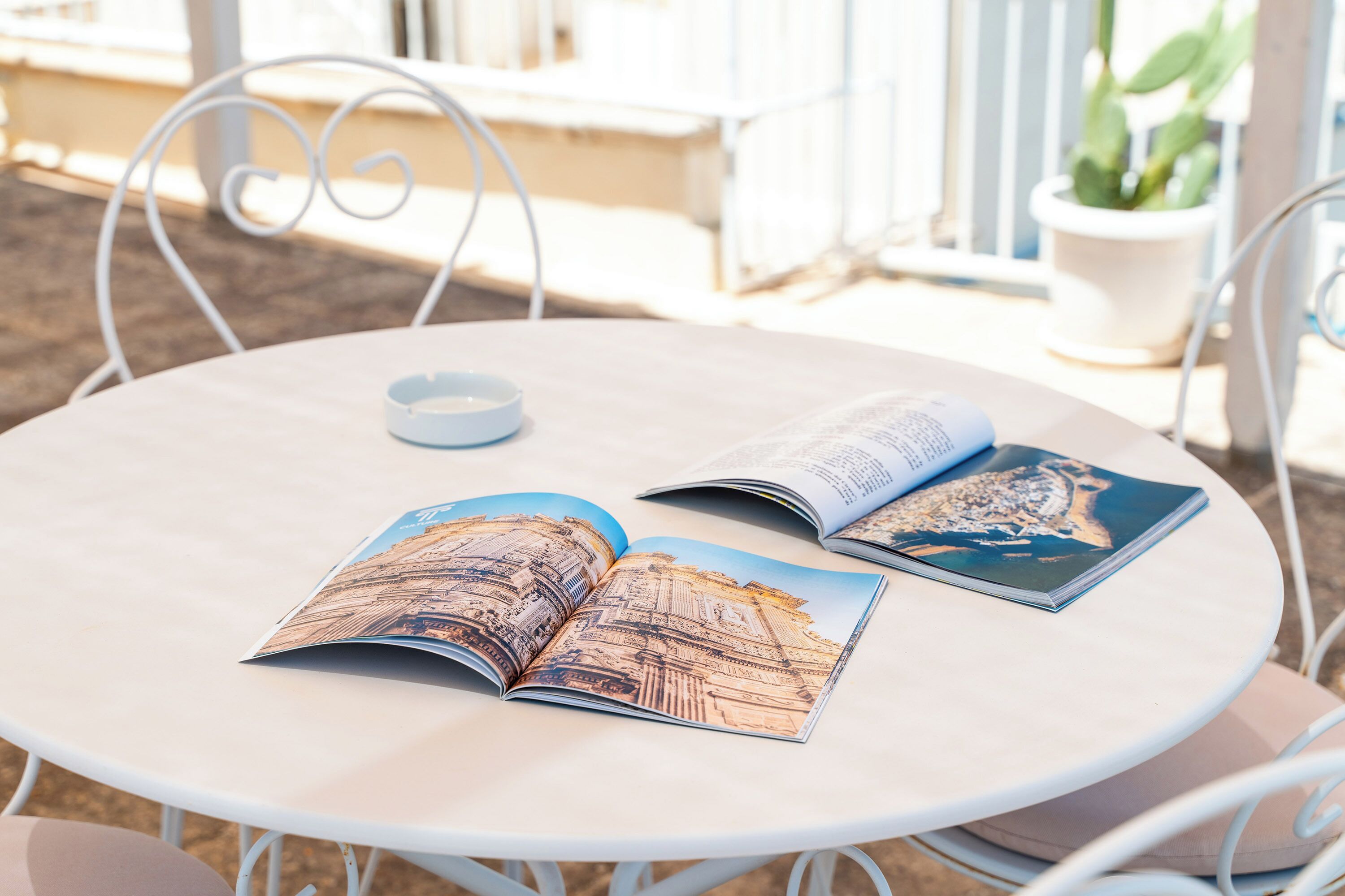 Une table ronde sur laquelle reposent deux magazines ouverts, un petit bol blanc et, au fond, une plante en pot.