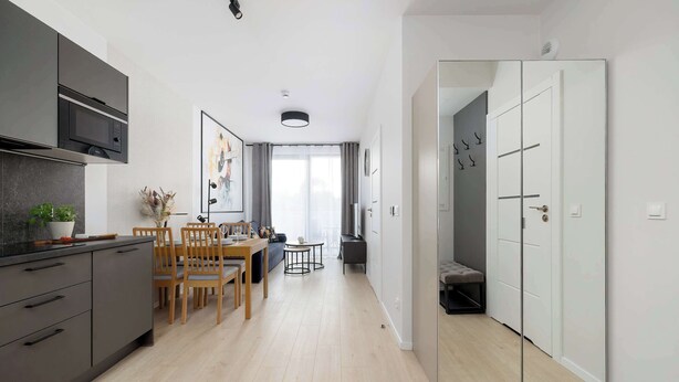 A modern kitchen with dark cabinets and a microwave, a dining area with wooden chairs and a table, and a hallway leading to a mirrored wardrobe. - Air Condtioned Apartment | Rooftop Pool (Mielno)