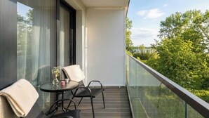 A balcony with a glass railing, a table with two chairs, and a view of trees. - Air Condtioned Apartment | Rooftop Pool (Mielno)