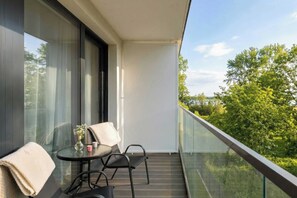 Ein Balkon mit Glasgeländer, ein Tisch und zwei Stühle sowie Blick auf Bäume.