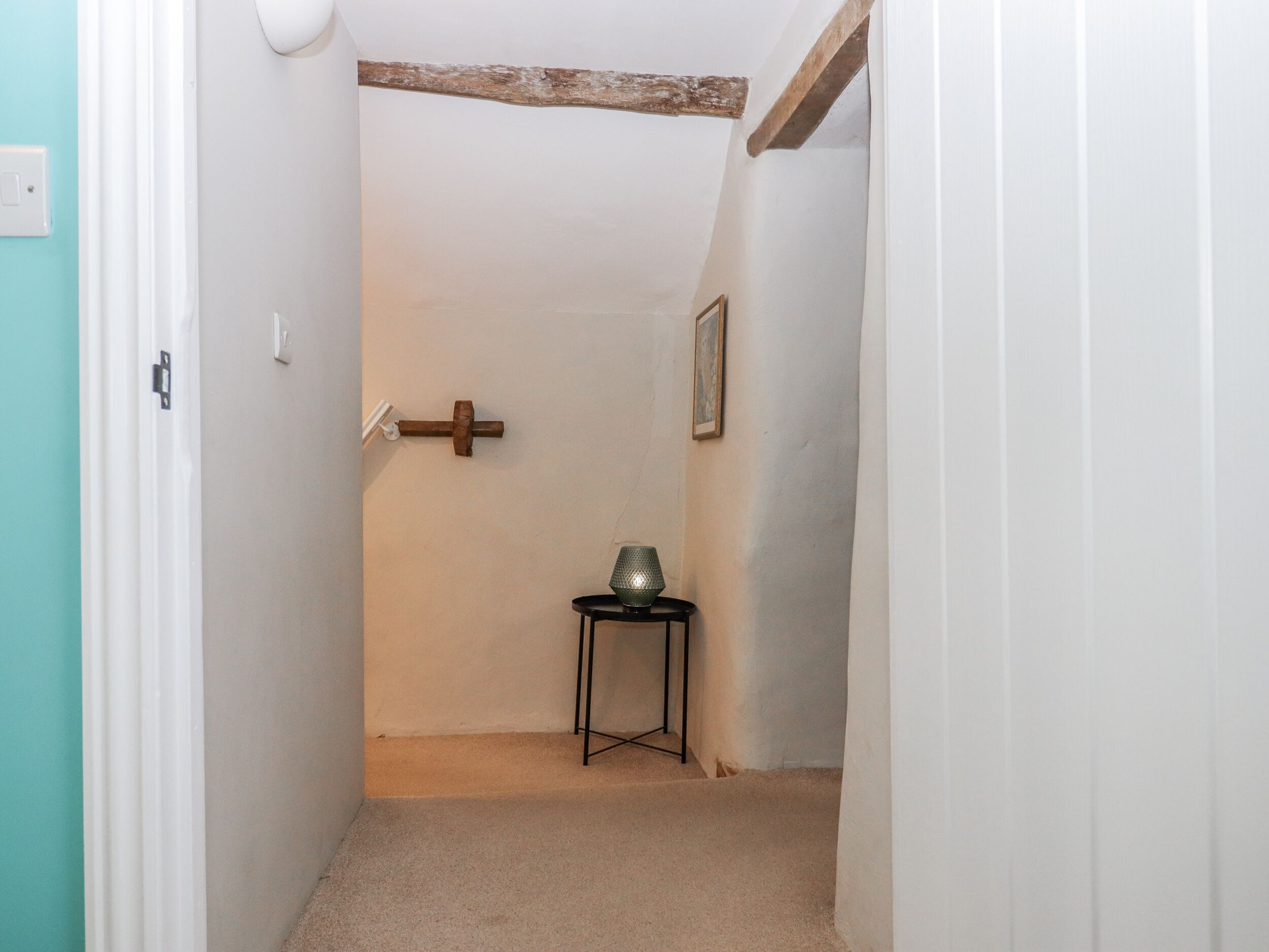 Un pasillo estrecho con techo de vigas de madera, una pequeña mesa con lámpara y una puerta con interruptor de luz.