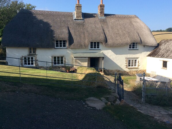 A traditional thatched-roof house with a stone chimney, a small porch, and a garden with a metal gate. - Westacombe Farmhouse (Exeter)