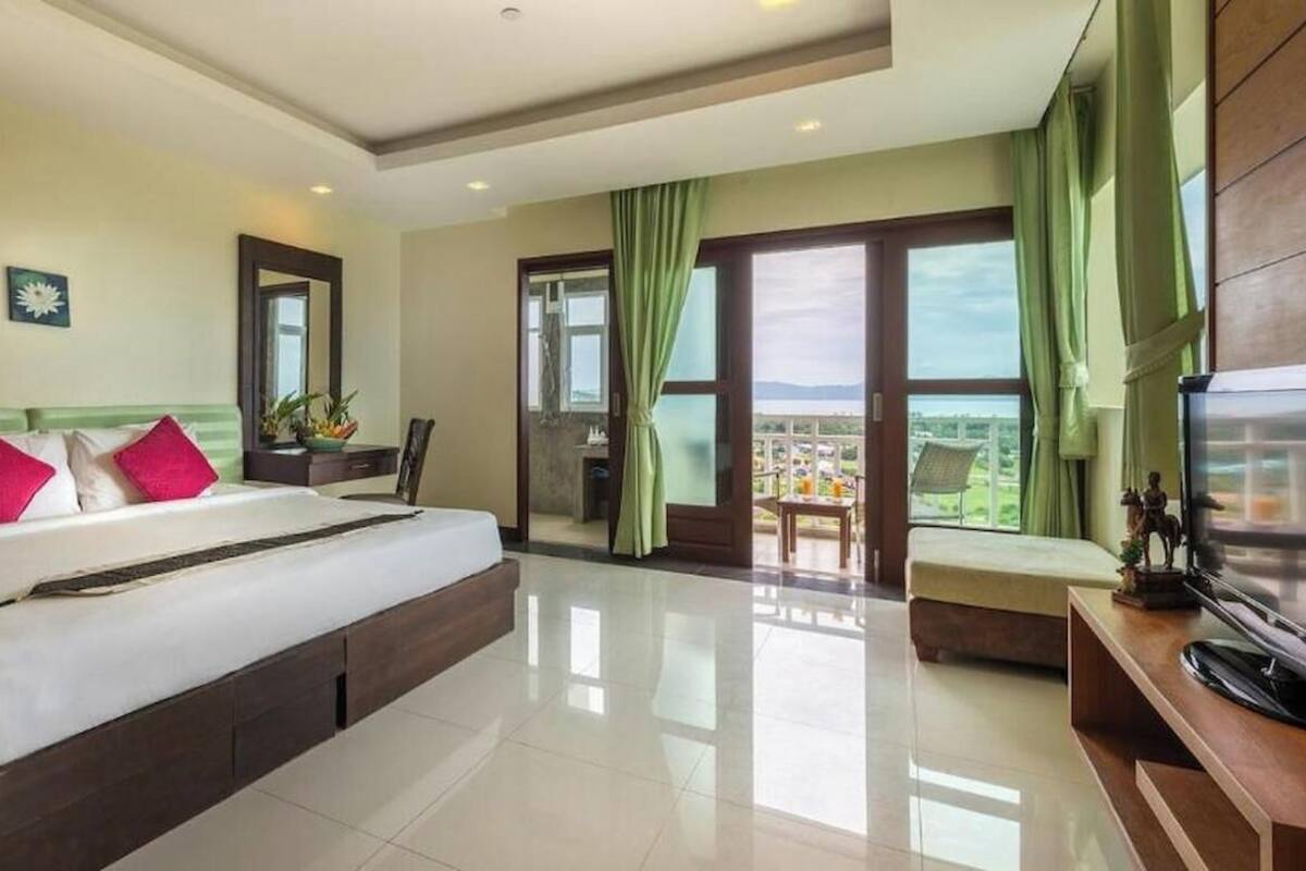 A hotel room with a large bed, a desk, a chair, a TV, and a balcony with a view.