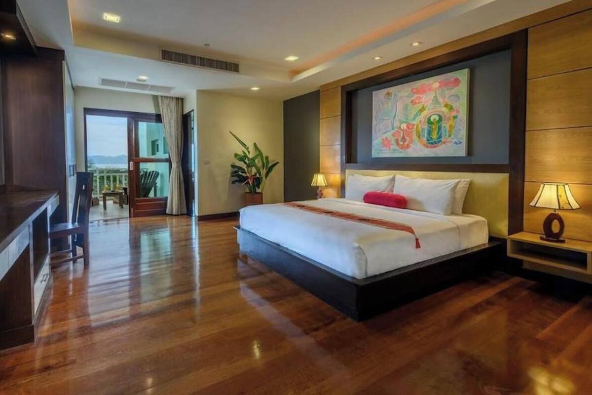 A hotel room with a bed, a desk, a TV, a plant, and a painting on the wall.