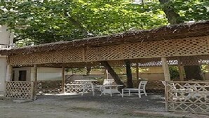 A covered outdoor seating area with tables and chairs, a building in the background, and a tree providing shade. - Palaash Bon Homestay (Guwahati)