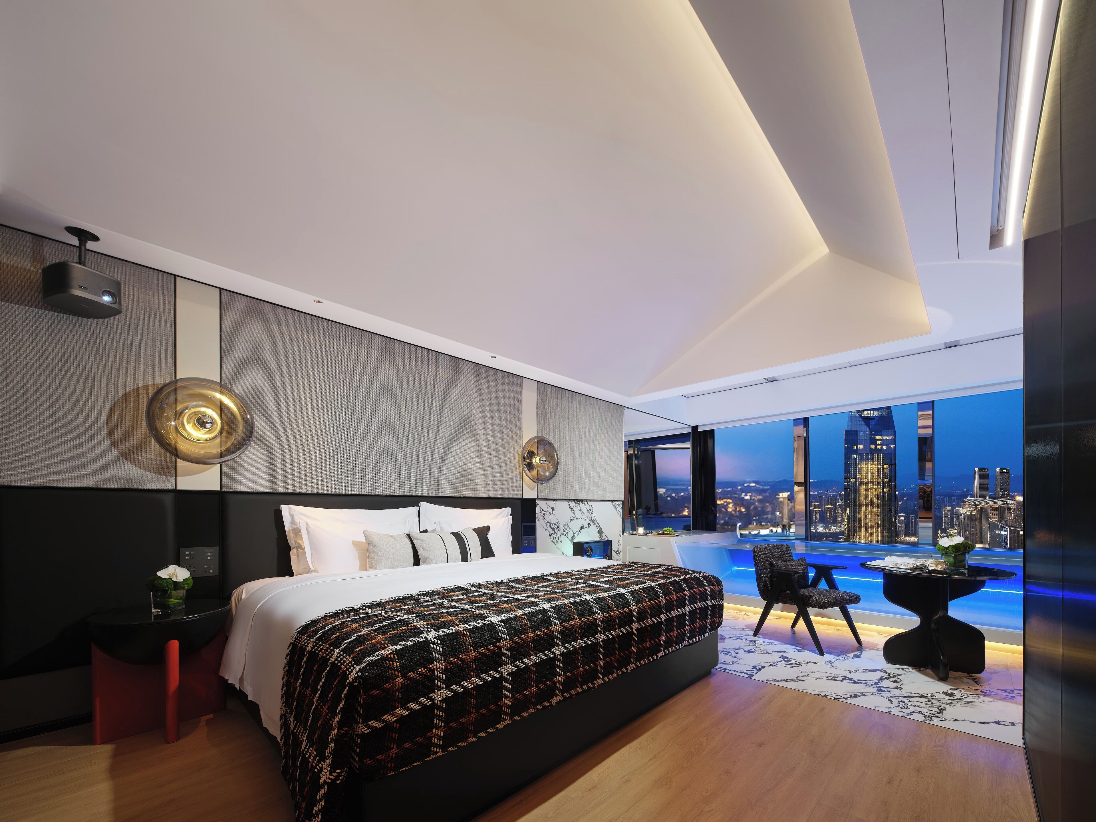 A modern bedroom with a large bed, a view of the cityscape, and a small table with a chair.