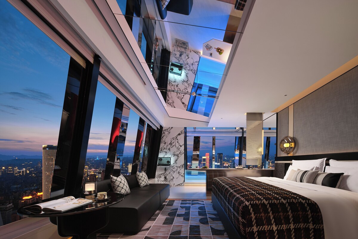A modern hotel room with a city view, a large bed, a sofa, a dining area, and a kitchen.