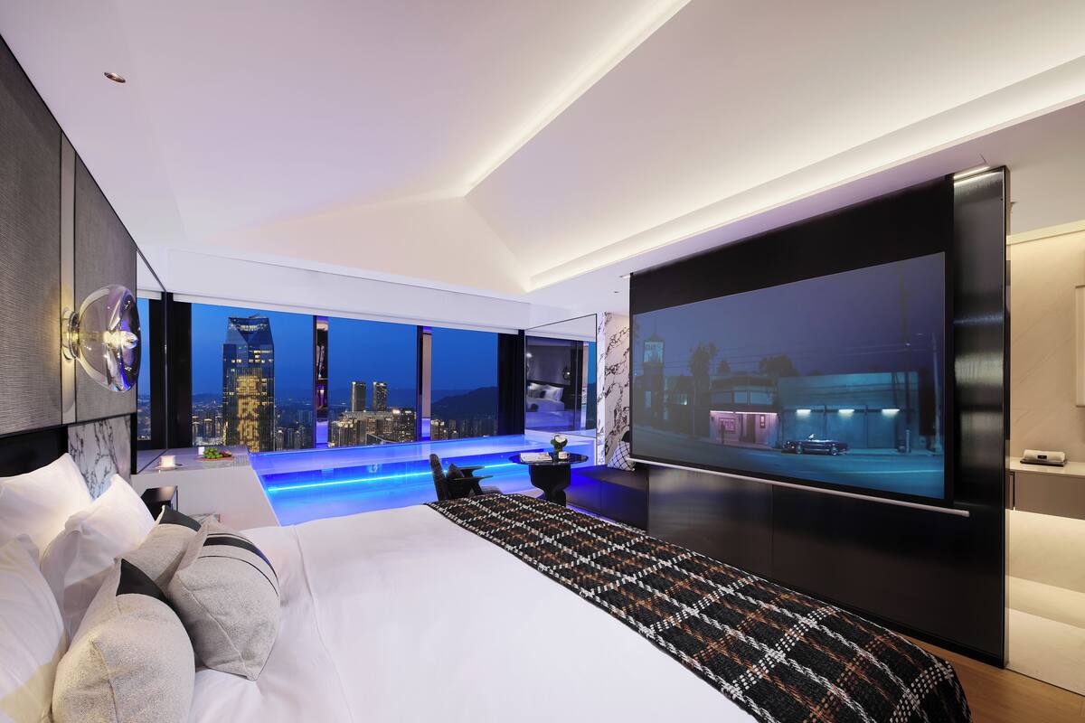 A modern hotel room with a city view, a large bed, a flat-screen TV, and a unique ceiling design.