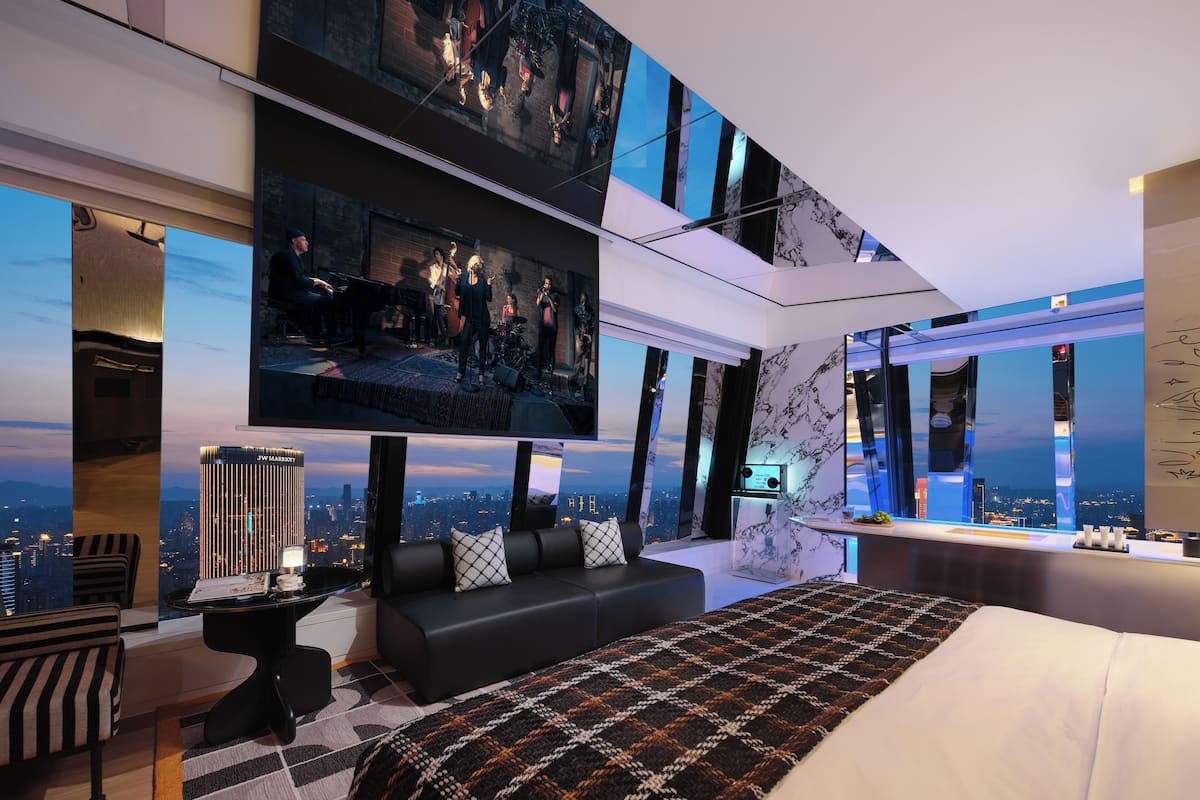 A modern hotel room with a large bed, a seating area, a TV screen, and a city view.