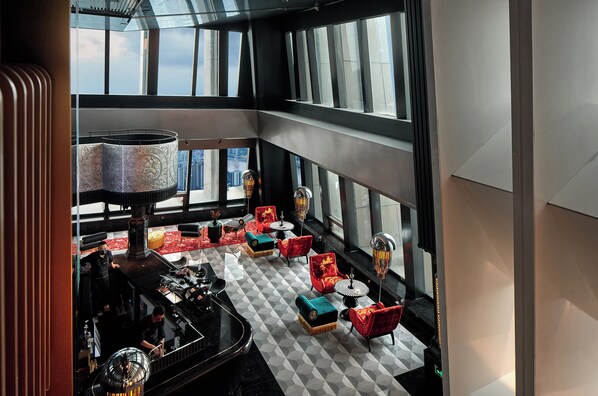 Vestíbulo de un hotel de gran altura con suelo a cuadros, asientos en rojo y negro, y amplias ventanas con vistas a la ciudad.