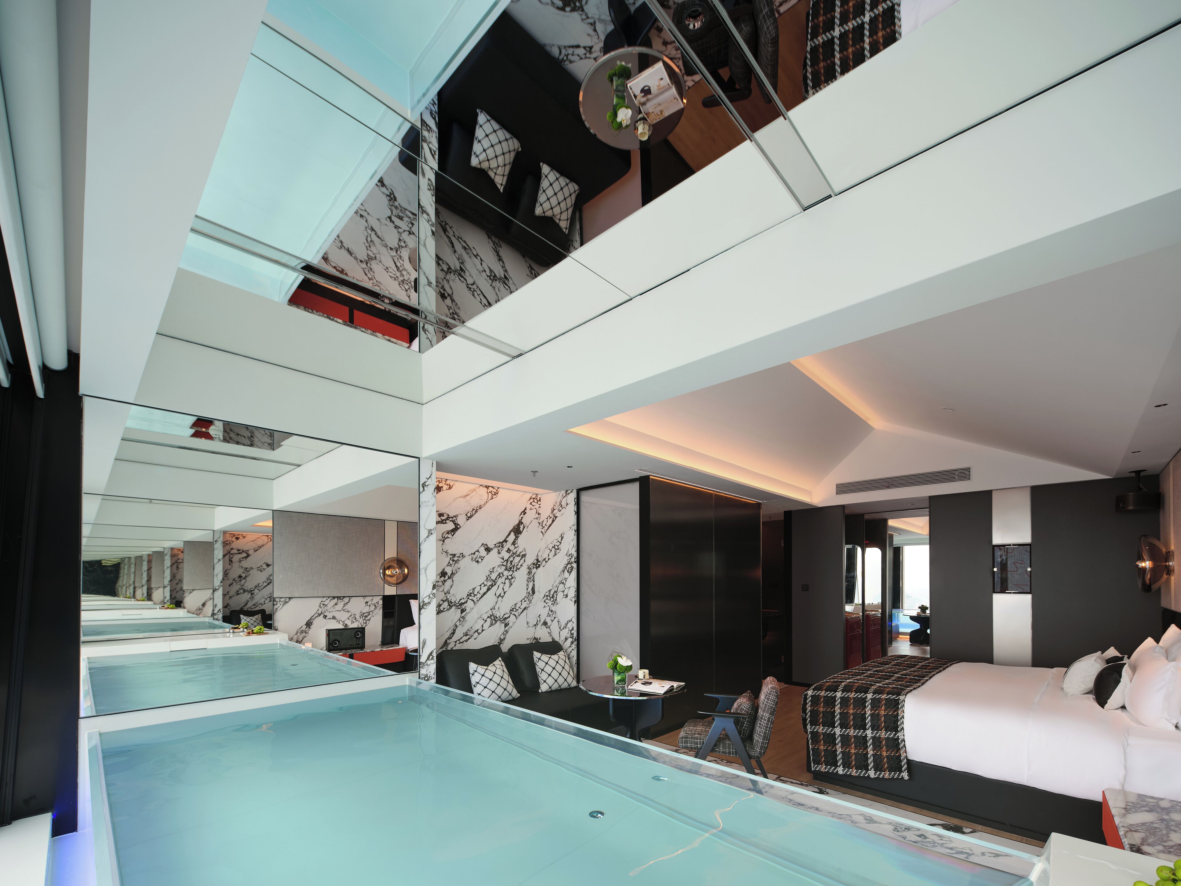 A modern hotel room with a glass-enclosed swimming pool, a bed, a seating area, and a view of the city.