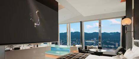 A modern hotel room with a large bed, a TV, a small table, and a view of mountains and a cityscape.
