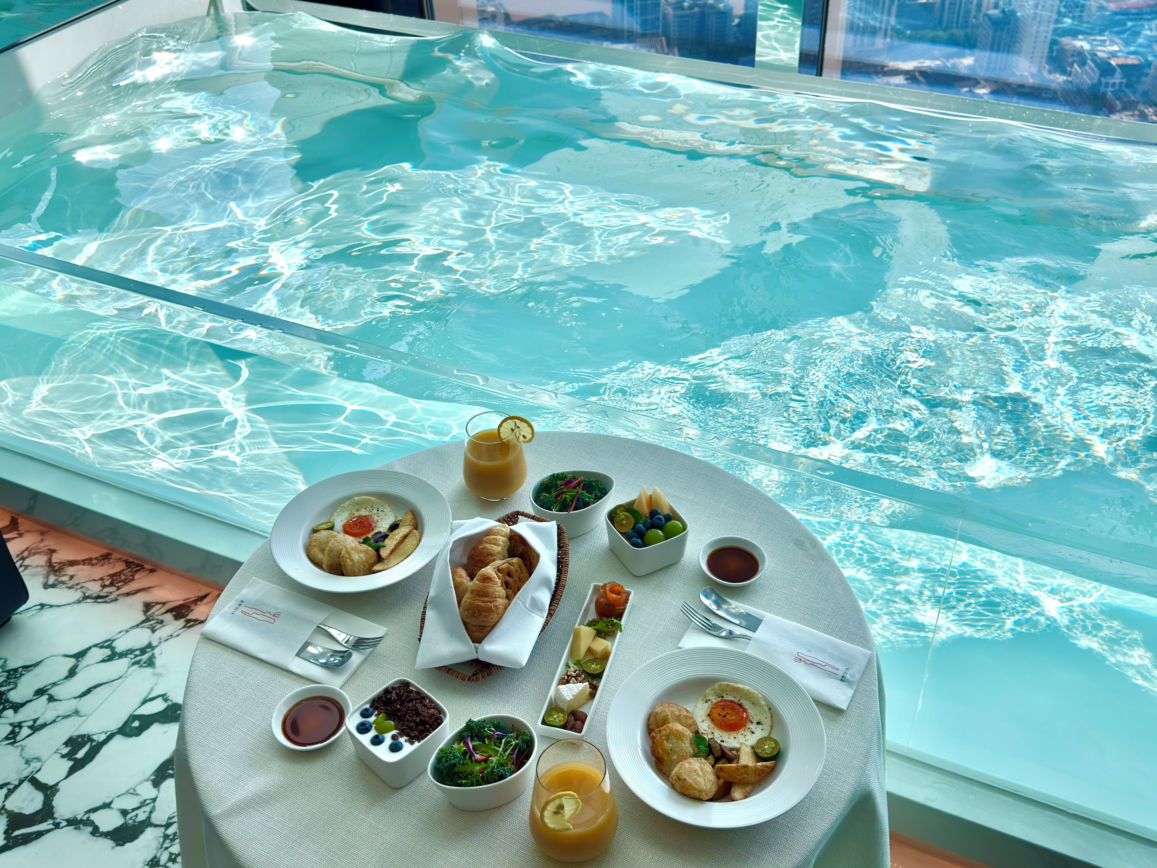 A dining table with breakfast items and a glass of orange juice, overlooking a pool.