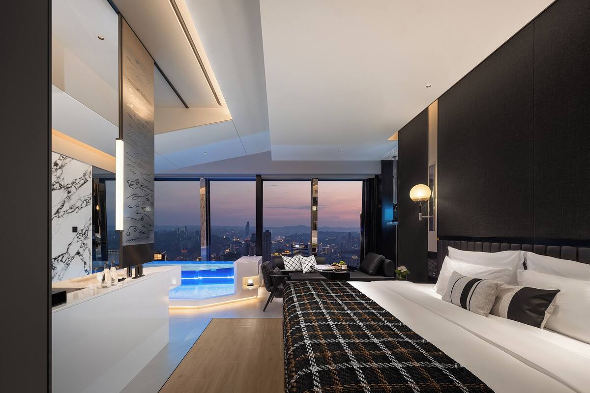 A modern hotel room with a large bed, a desk, a view of the city, and a small pool.