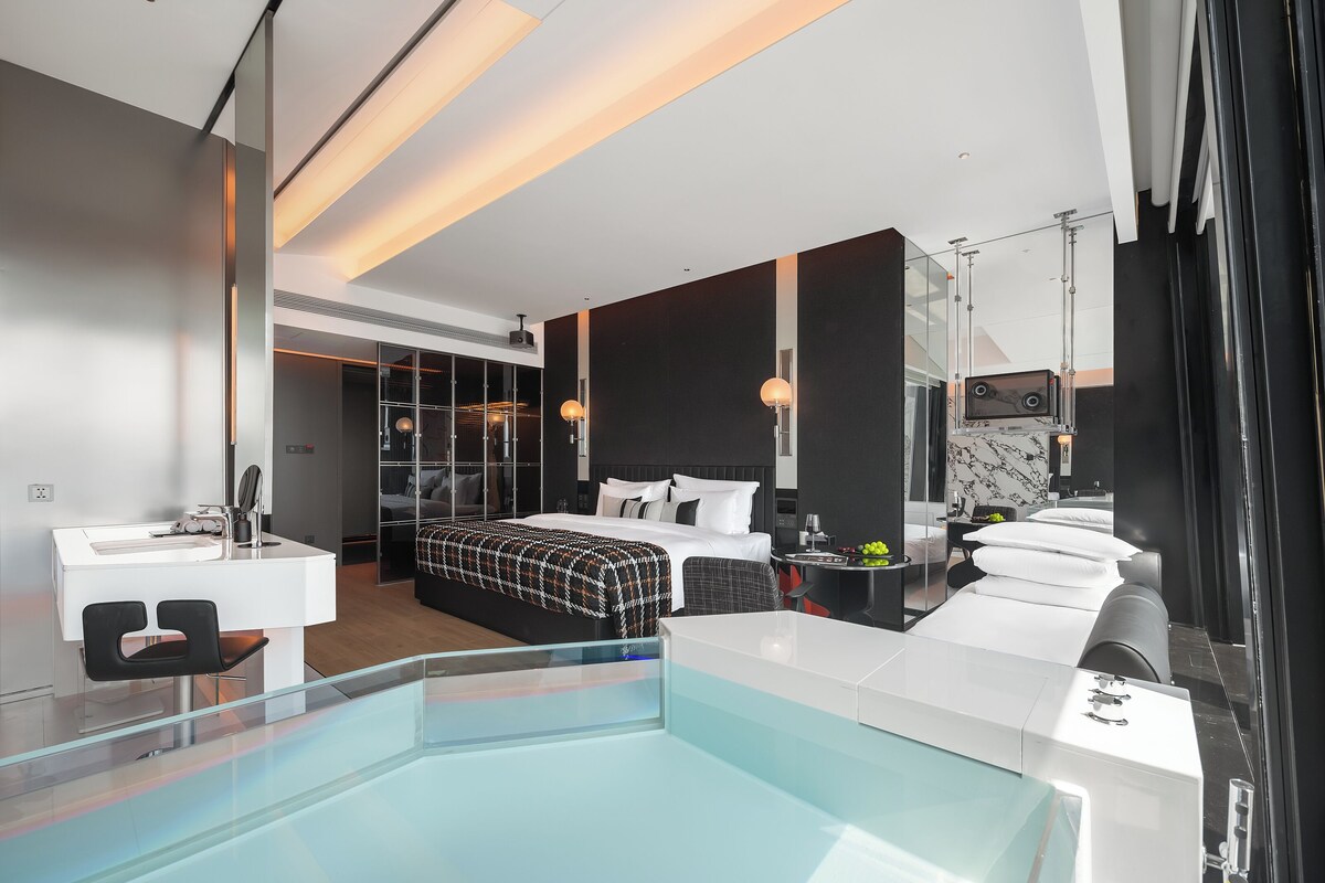 A modern hotel room with a large bed, a glass-enclosed walk-in closet, a separate bathroom, and a pool area.