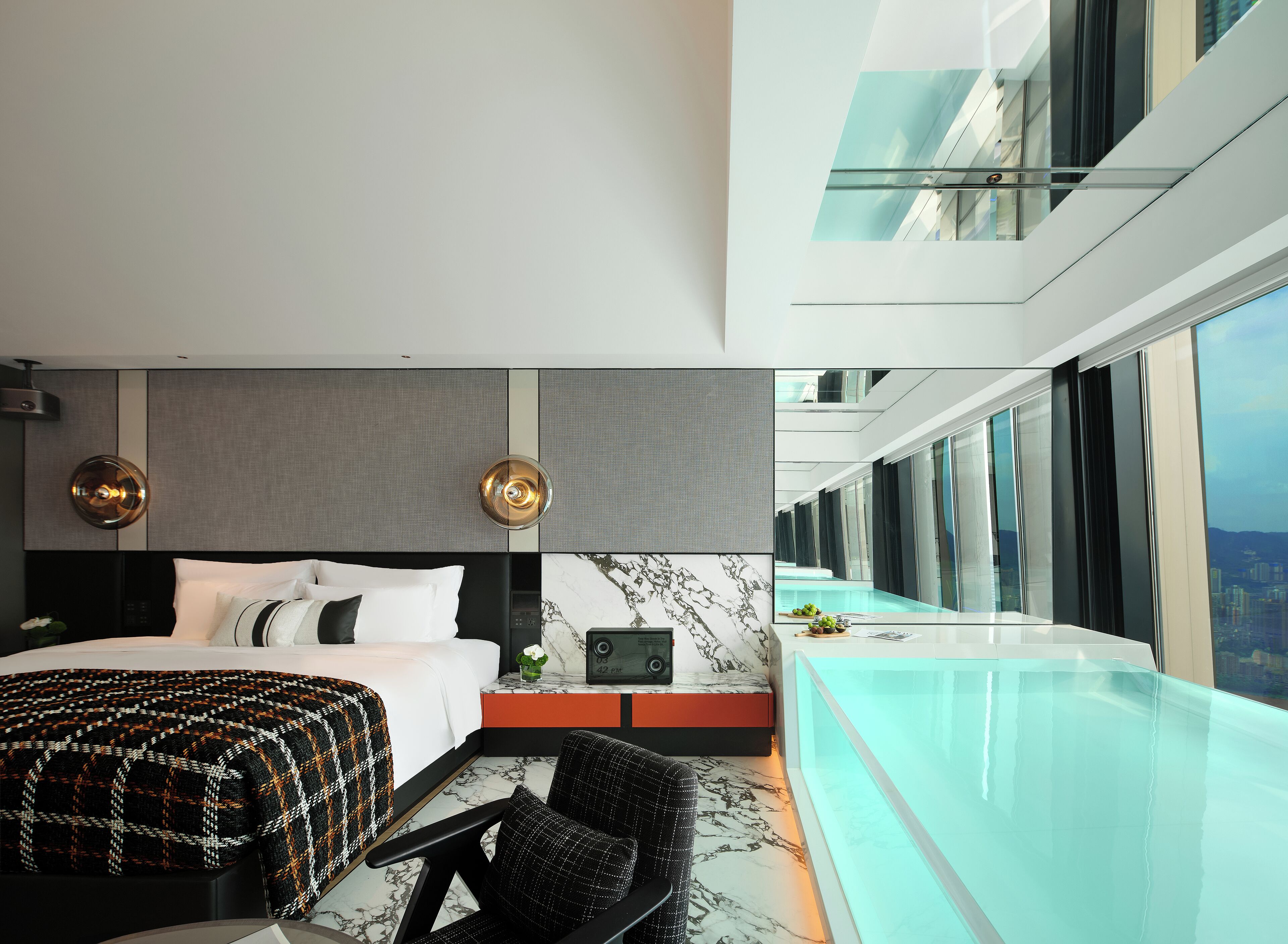 A modern hotel room with a large bed, a seating area, a small table, a laptop, and a view of a cityscape through large windows.