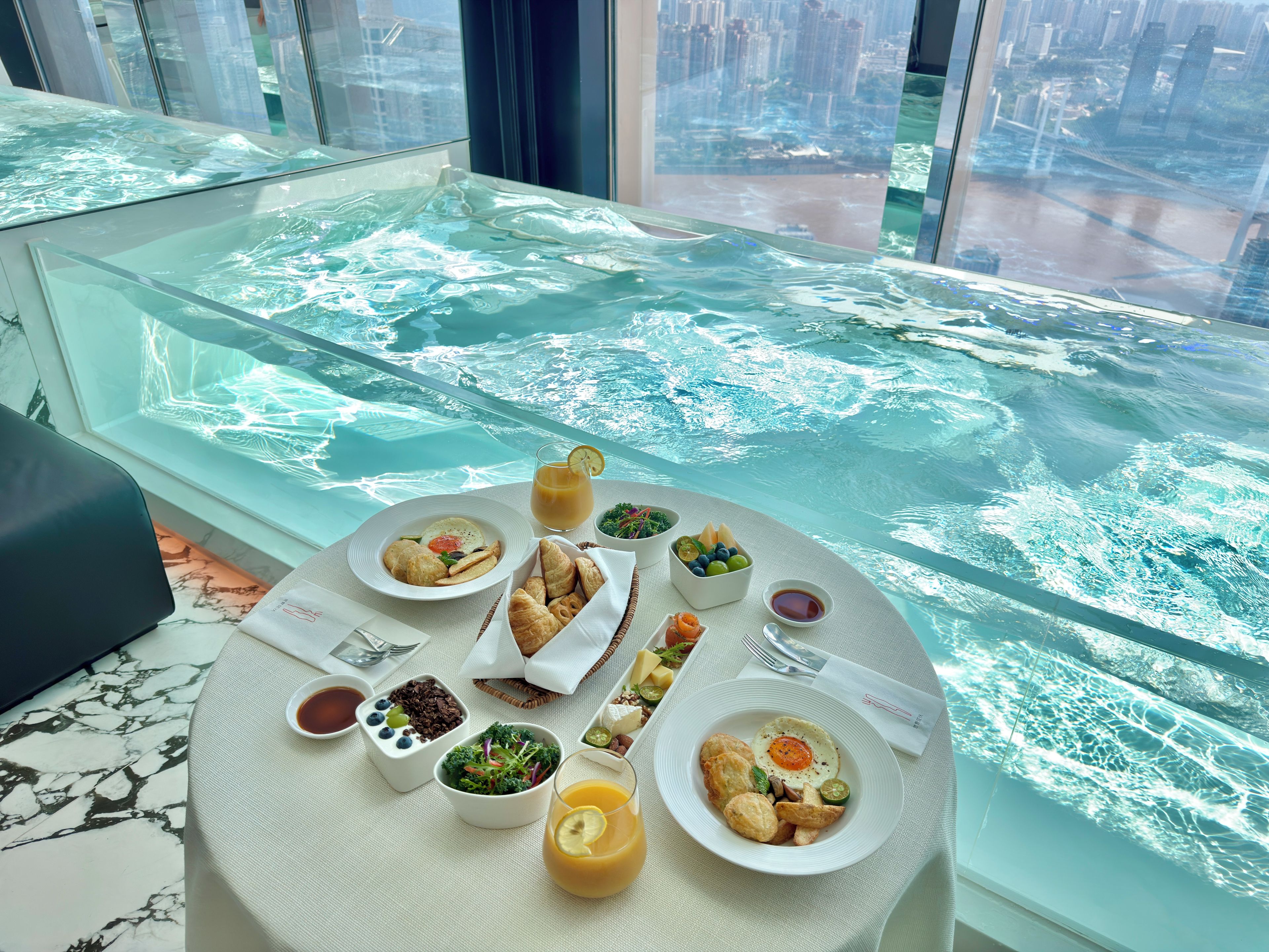 Una mesa de comedor con alimentos para el desayuno y una mesa de vidrio con vista a un paisaje urbano.
