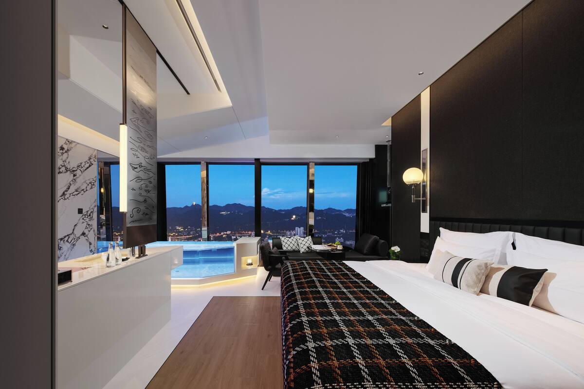 A modern hotel room with a large bed, a desk, a view of the cityscape, and a jacuzzi.