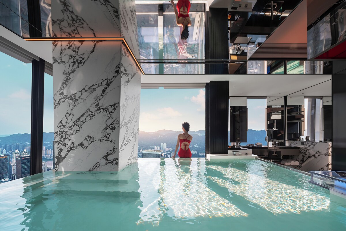 A modern indoor infinity pool with a city view, marble walls, and a red swimsuit.