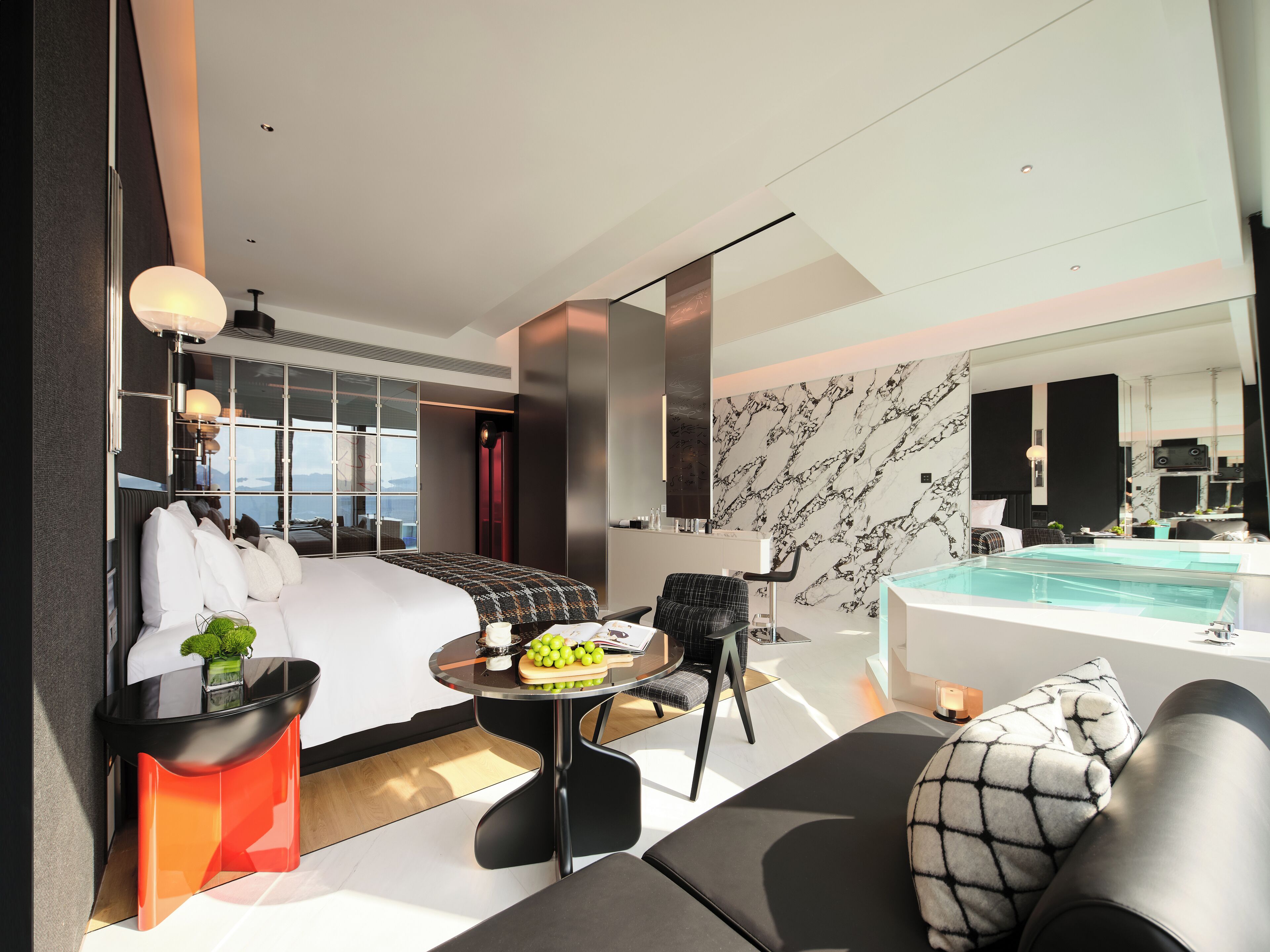 A modern hotel room with a bed, a round table with a fruit bowl, a sofa, a small round table, a large mirror, and a jacuzzi.