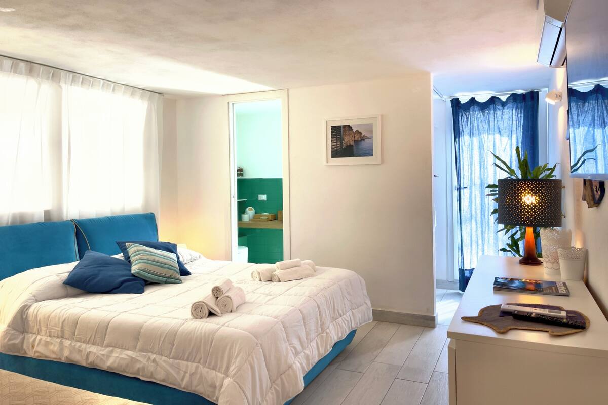 A bedroom with a bed, a bedside table with a lamp, a window with curtains, and a green wall with a picture.