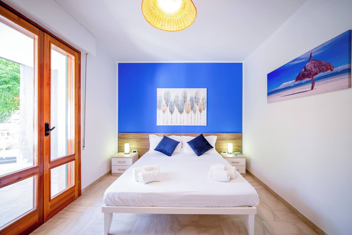 A hotel room with a large bed, two bedside tables, a painting on the wall, and a view through a large window.