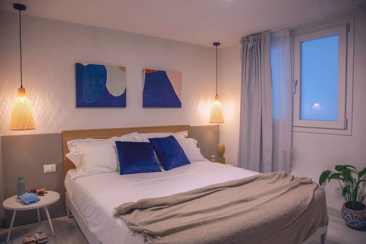 A neatly made bed with white and blue bedding, two wall-mounted lamps, a window with curtains, and a small table with a water bottle and slippers.