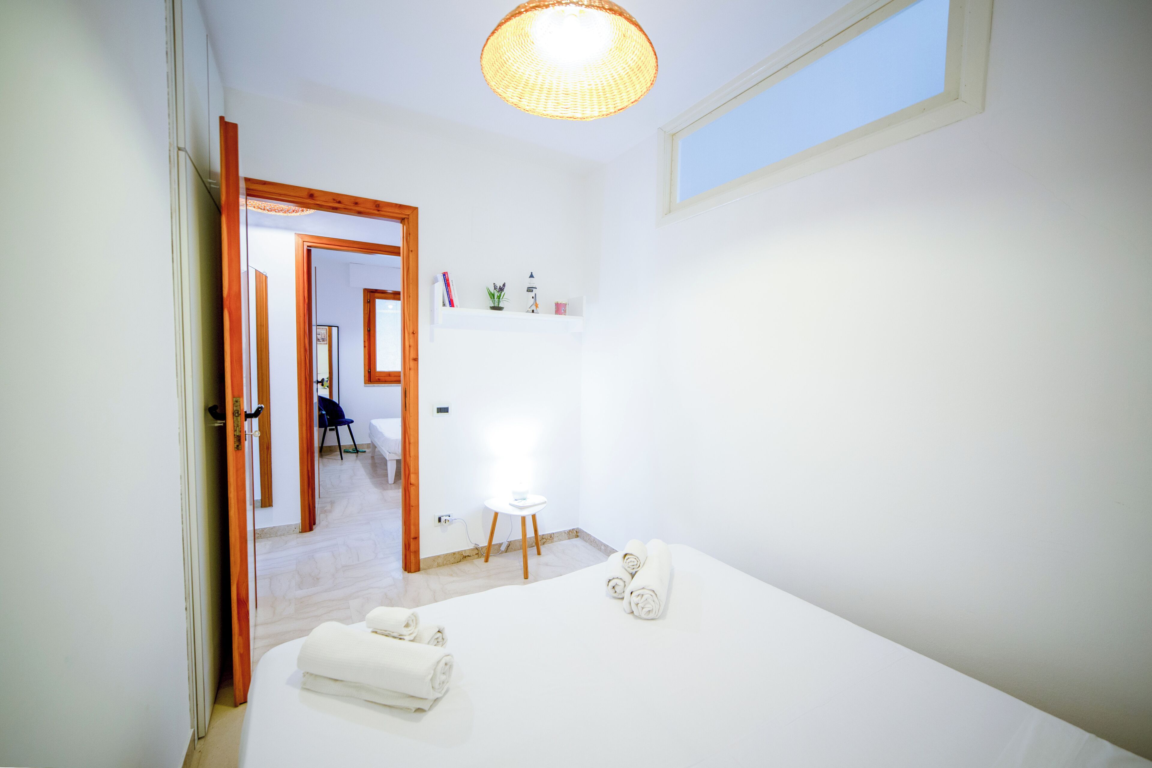Habitación de hotel minimalista con cama, una mesita de noche, una silla, un escritorio y una estantería con objetos decorativos.