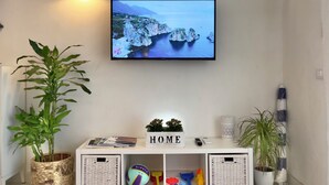 A living room with a flat-screen TV displaying a coastal scene, a white console table with a 'HOME' sign, and two potted plants. - Marina Beach Apartments (Alcamo)