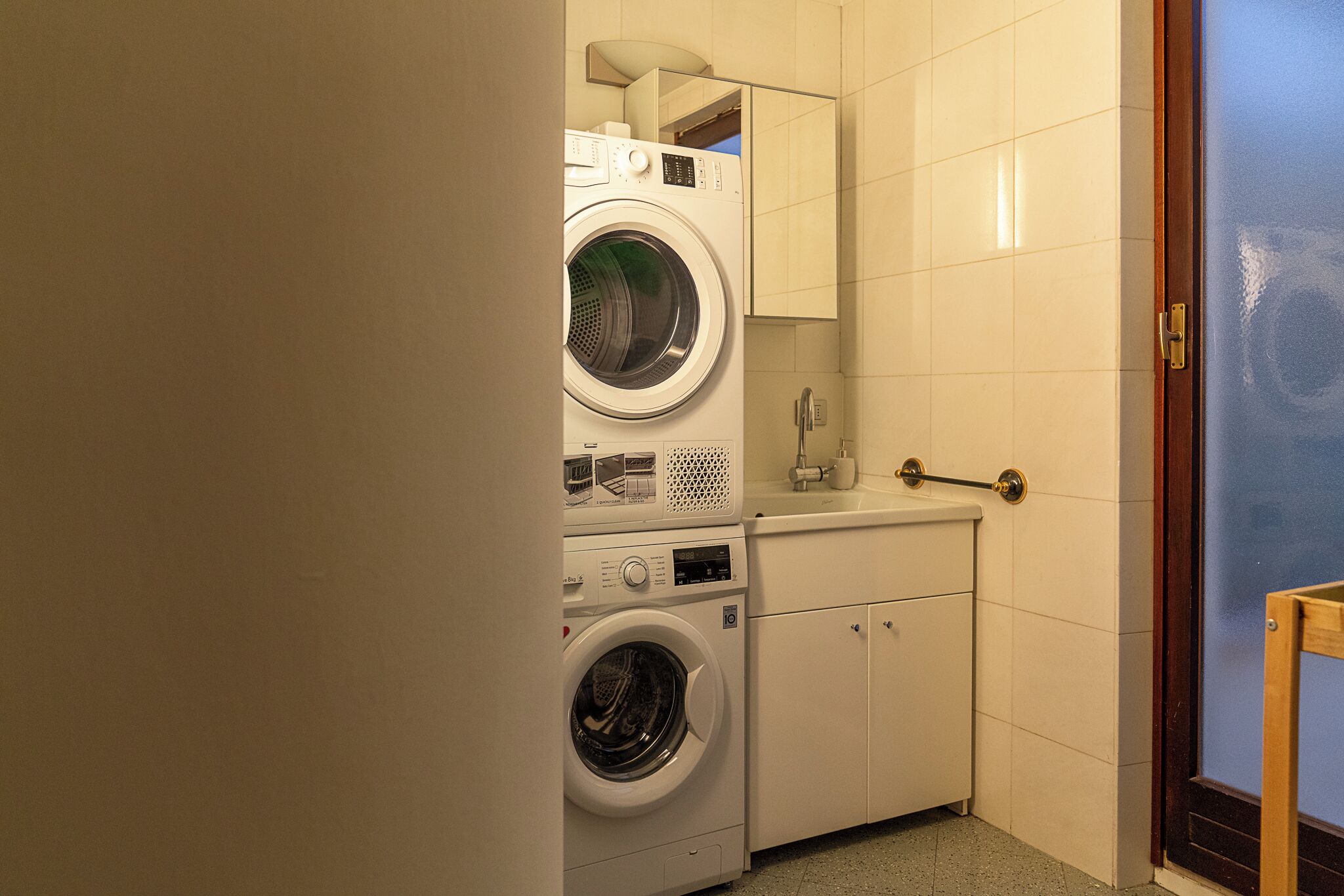 Une petite buanderie équipée d’un lave-linge et d’un sèche-linge à chargement frontal, d’un petit plan de travail avec un miroir et d’une porte dotée d’une poignée.