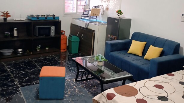 Een knus woongedeelte met een blauwe bank, een klein bijzettafeltje, een keuken met een spoelbak en kastjes, en een raam dat natuurlijk licht laat binnen.